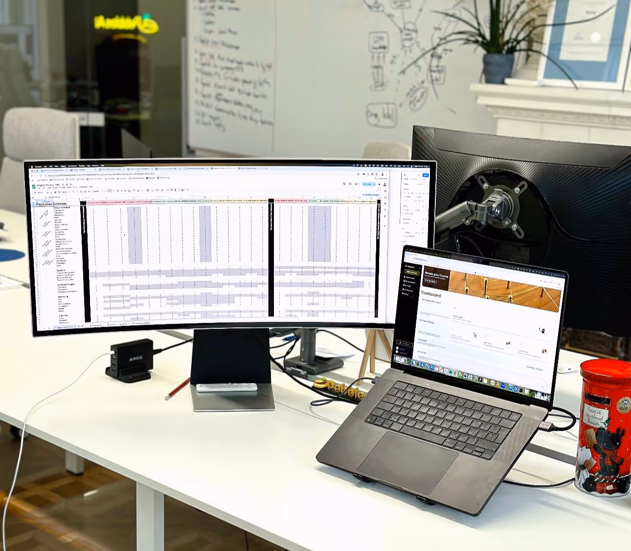 An office desk with a laptop, wide monitor and notes on the wall.